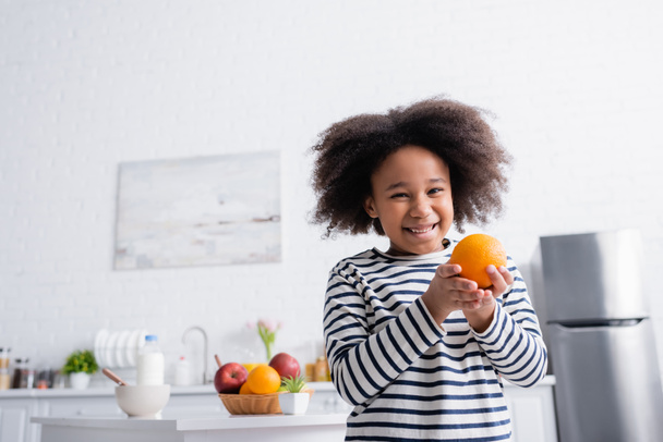 smiling african american kid holding orange near breakfast on kitchen table - Photo, Image