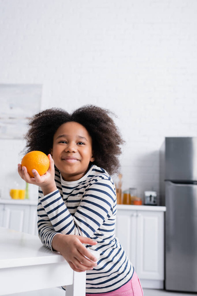 feliz afroamericano chica sosteniendo maduro naranja mientras de pie cerca de mesa en cocina - Foto, imagen