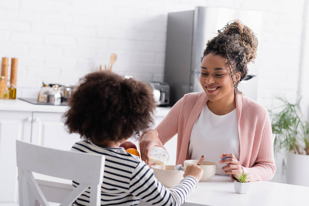 smiling african american woman pouring milk during breakfast with daughter - Photo, Image