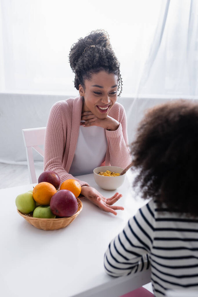 アフリカ系アメリカ人の幸せな女性が朝食中に娘と話をしながらジェスチャーをしながら - 写真・画像