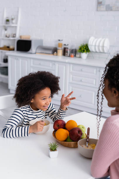 アフリカ系アメリカ人の少女が朝ご飯を食べながら前景のぼやけた母親と話をしながら - 写真・画像
