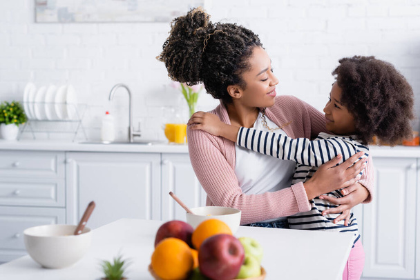 alegre afroamericana madre e hija abrazando en cocina cerca de frutas frescas en primer plano borrosa - Foto, imagen