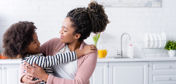 feliz afro-americana mãe e filha olhando um para o outro enquanto abraçando na cozinha, bandeira - Foto, Imagem