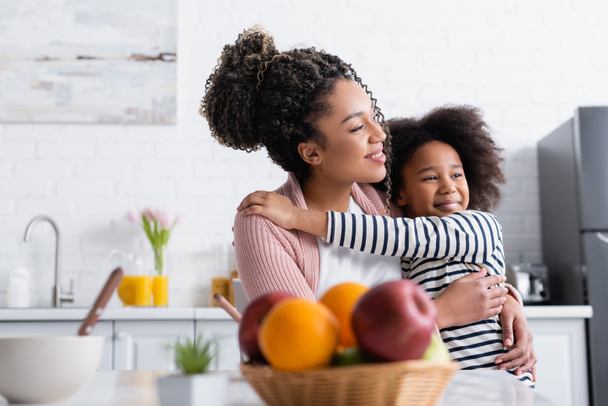 femme afro-américaine gaie et enfant embrassant tout en regardant loin près des fruits sur le premier plan flou - Photo, image