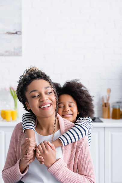 heureux afro-américaine fille avec les yeux fermés tenant la main de la mère gaie dans la cuisine - Photo, image