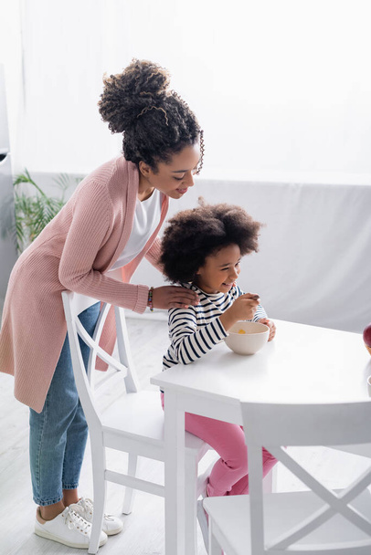 sorrindo mulher afro-americana tocando ombros de filha tomando café da manhã na cozinha - Foto, Imagem