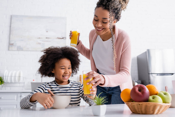 smiling african american woman giving orange juice to daughter near fruits on blurred foreground - Photo, Image