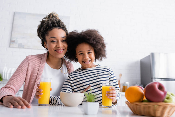 alegre africano americano madre y hija mirando cámara cerca naranja jugo y frutas en borrosa primer plano - Foto, Imagen