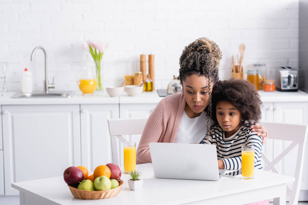 elképedt afro-amerikai anya és lánya használ laptop közelében narancslé és gyümölcsök fonott kosárban - Fotó, kép