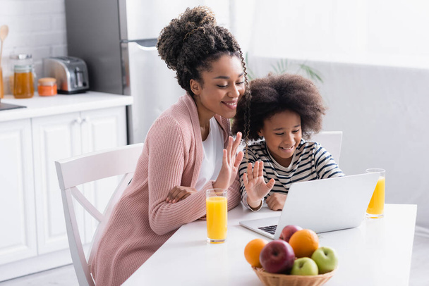 enfant afro-américain joyeux avec la mère agitant les mains lors d'un appel vidéo sur un ordinateur portable dans la cuisine - Photo, image