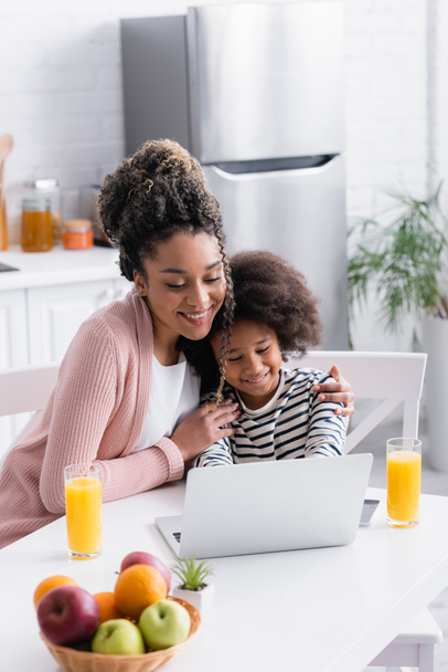 Gülümseyen Afrikalı Amerikalı anne, mutfakta dizüstü bilgisayarla kızını kucaklıyor. - Fotoğraf, Görsel