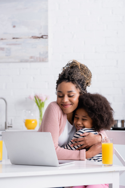 happy african american mom embracing daughter while watching film on laptop during breakfast - Photo, Image