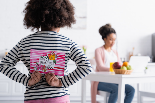 вид на африканского американского ребенка, держащего счастливую открытку на день матери рядом с мамой, работающей на кухне на размытом фоне - Фото, изображение