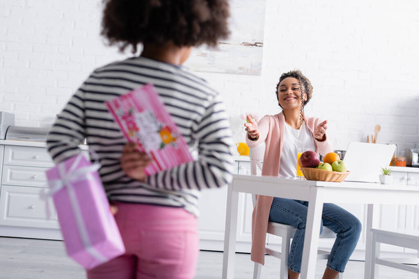 Neşeli afro-amerikan kadını kızının yanına uzanan hediye ve bulanık ön planda mutlu anneler günü kartı. - Fotoğraf, Görsel