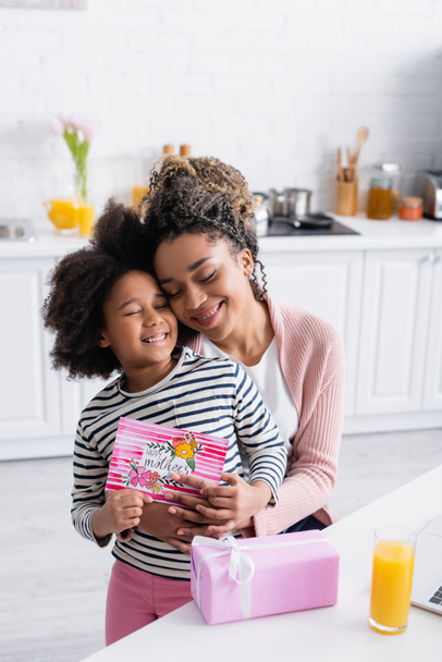 feliz africana americana mujer abrazando hija celebración feliz madres día tarjeta cerca de caja de regalo - Foto, Imagen