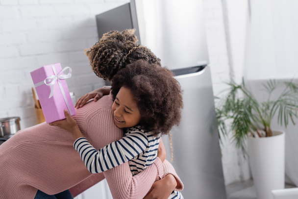 Αφροαμερικανή που αγκαλιάζει χαρούμενη κόρη κρατώντας δώρο για την γιορτή της μητέρας. - Φωτογραφία, εικόνα