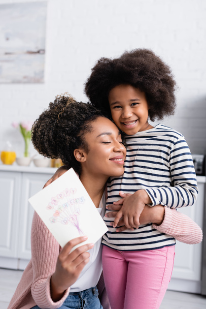 cheerful african american woman with closed eyes holding happy mothers day card while hugging smiling daughter - Photo, Image