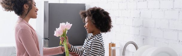 vista lateral da criança afro-americana apresentando tulipas frescas para mamãe, banner - Foto, Imagem