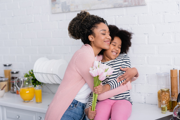 Afroamerikanerin umarmt Tochter, die mit Tulpen auf Küchentisch sitzt - Foto, Bild