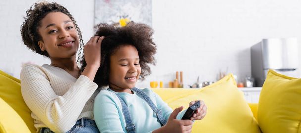 feliz afroamericana mujer tocando el pelo de hija sosteniendo mando a distancia mientras mira la televisión, pancarta - Foto, Imagen