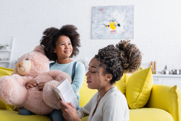 若いアフリカ系アメリカ人女性がテディベアとソファに座っている子供に本を読んで - 写真・画像