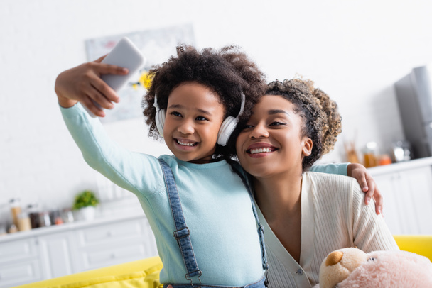 alegre africana americana chica en auriculares tomando selfie en smartphone con feliz madre - Foto, imagen