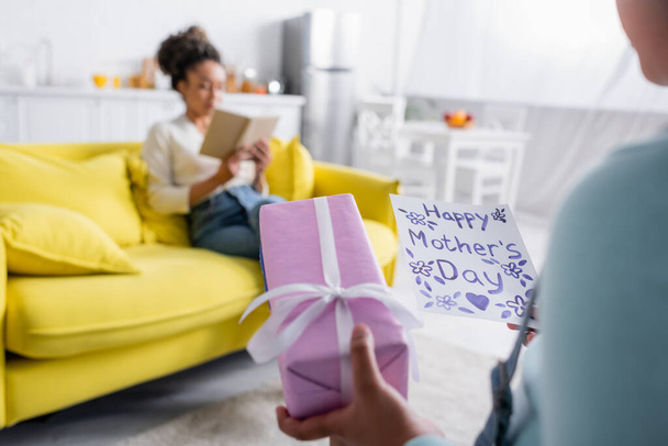 niño sosteniendo feliz madres tarjeta de día cerca de la mujer afroamericana leyendo libro en el sofá, fondo borroso - Foto, imagen