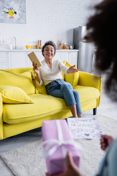 feliz mujer afroamericana sentada en un sofá con la mano extendida cerca de la hija con tarjeta de felicitación y regalo en primer plano borroso - Foto, imagen