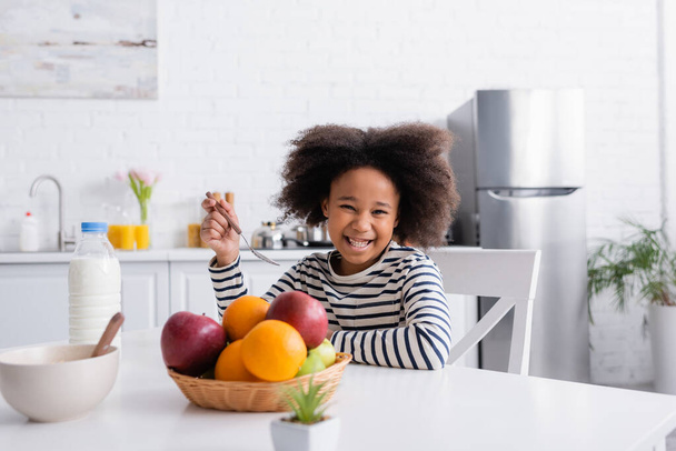 niño afroamericano complacido sosteniendo cuchara cerca de la leche y las frutas, borrosa primer plano - Foto, Imagen