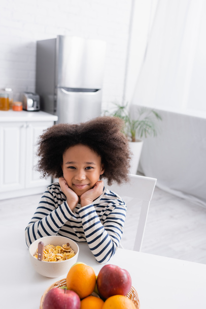 Радостная африканская американка сидит рядом с кукурузными хлопьями и свежими фруктами на кухонном столе - Фото, изображение