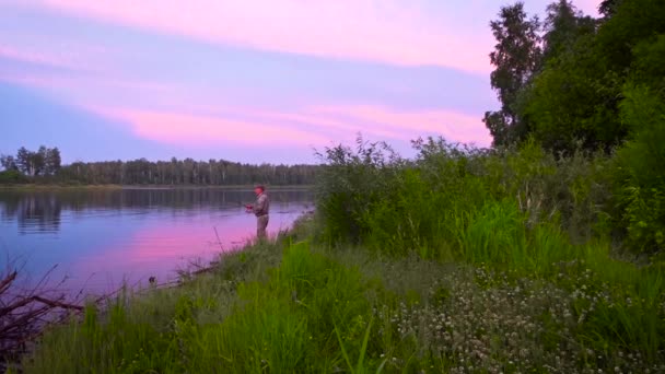 De visser gooit een draaiende hengel - hij is aan het vissen. Zonsondergang, roze wolken boven het meer. Zomer landschap. - Video