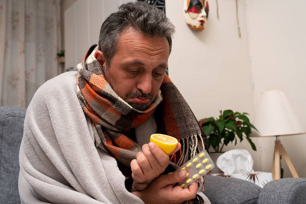 Hombre adulto enfermo sintiéndose mal usando bufanda haciendo expresión triste sosteniendo medicina mirando limón para la inmunidad vitaminc como concepto pandémico del virus de la gripe fría - Foto, Imagen