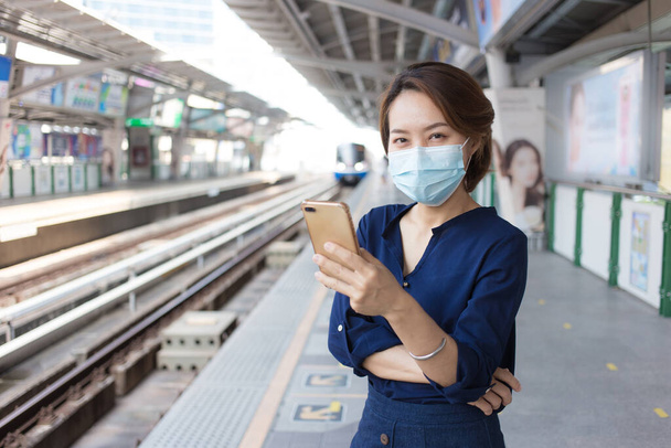 Asiatin mit kurzen Haaren spielt Smartphone beim Warten auf Zug am Bahnhof und trägt medizinische Gesichtsmaske als neuen normalen Lebensstil. - Foto, Bild