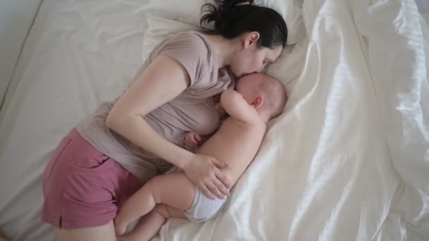 Fiatal anya fekszik újszülött aranyos csecsemő meztelen kisfiú az ágyban, tartja őt a karját, ölelés és szoptat anyatejjel. Egészséges gyermek, a kórház és a boldog anyaság fogalma. Gyermekszoba - Felvétel, videó