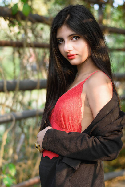Portret bardzo pięknej, atrakcyjnej Indianki ubranej w czerwony strój z czarną kurtką, pozującej przed tropikalnymi zielonymi liśćmi w słoneczny dzień w rozmazanym zielonym tle. Styl życia i moda. - Zdjęcie, obraz