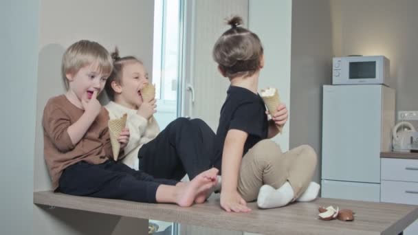 Veljet ystävät lapset istuvat pöydällä ja syövät jäätelöä yhdessä kotona keittiössä. - Materiaali, video