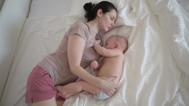 Fáradt anya alszik újszülött aranyos csecsemő meztelen kisfiú az ágyban, tartja őt a karját, ölelés és szoptat anyatejjel. Egészséges gyermek, a kórház és a boldog anyaság fogalma. Gyermekszoba - Felvétel, videó