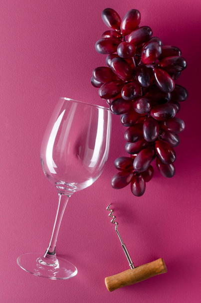 Immagine verticale di uva rossa, vino di vetro e cavatappi manico sulla superficie rosa - Foto, immagini