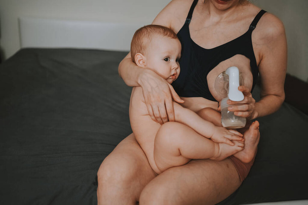 Young mother with newborn cute infant naked baby boy, holding him on arms, using breast pump hugging and breastfeeds with breast milk. Healthy child, concept of hospital and happy motherhood. Nursery. - Photo, Image