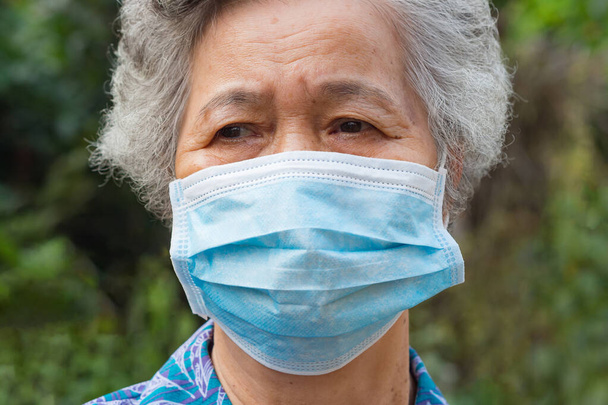 Porträt einer Seniorin, die eine Gesichtsmaske trägt und wegschaut, während sie in einem Garten steht. Maske zum Schutz von Covid-19, Virus, Coronavirus usw. Konzept für ältere Menschen und Gesundheitsfürsorge. - Foto, Bild