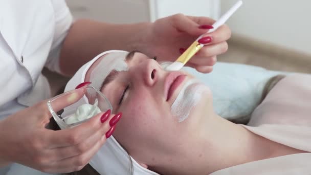 Cosmetólogo aplica máscara de arcilla con espátula en la cara de la mujer. Piel facial y tratamiento antienvejecimiento. Cosmetología y cuidado profesional de la piel facial. - Imágenes, Vídeo