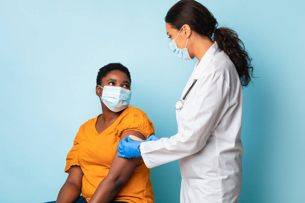 Ιατρικός εργαζόμενος εμβολιάζοντας μαύρη γυναίκα φορώντας μάσκες προσώπου, μπλε φόντο - Φωτογραφία, εικόνα