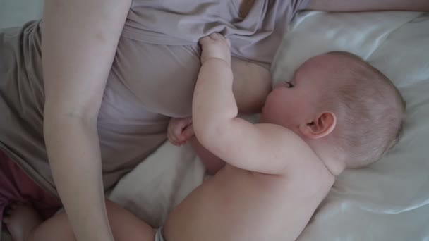 Fiatal anya fekszik újszülött aranyos csecsemő meztelen kisfiú az ágyban, tartja őt a karját, ölelés és szoptat anyatejjel. Egészséges gyermek, a kórház és a boldog anyaság fogalma. Gyermekszoba - Felvétel, videó