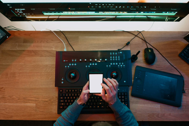 Editor de Vídeo Masculino sentado em seu computador usando tela em branco do telefone - Foto, Imagem