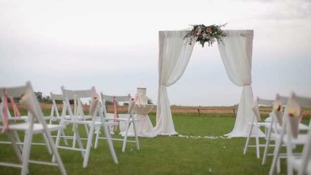 Łuk ślubny i puste krzesła ozdobione białymi i różowymi kwiatami. Atrybuty dekoracji ślubnej - Materiał filmowy, wideo