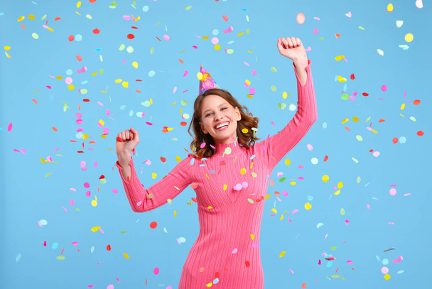 Femme optimiste en chapeau de fête souriant et dansant sous les confettis colorés tombant pendant la célébration de l'anniversaire sur fond bleu - Photo, image