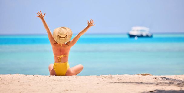 Vista posterior de la mujer en traje de baño vívido y sombrero de paja levantando brazos mientras está sentado en la orilla arenosa del océano turquesa - Foto, imagen