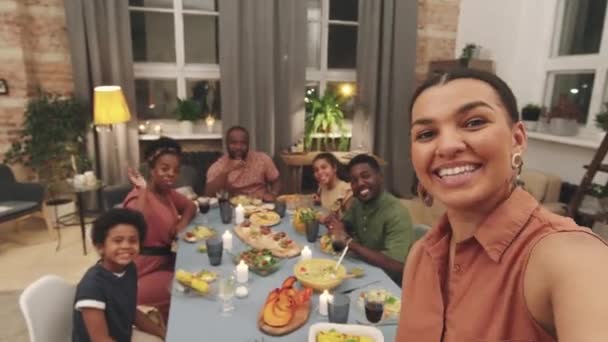 Cintura palmare di attraente donna di razza mista che si filma mentre cena con una grande famiglia afro seduta a tavola in soggiorno. Tutti si divertono insieme, ridendo e salutando alla telecamera - Filmati, video