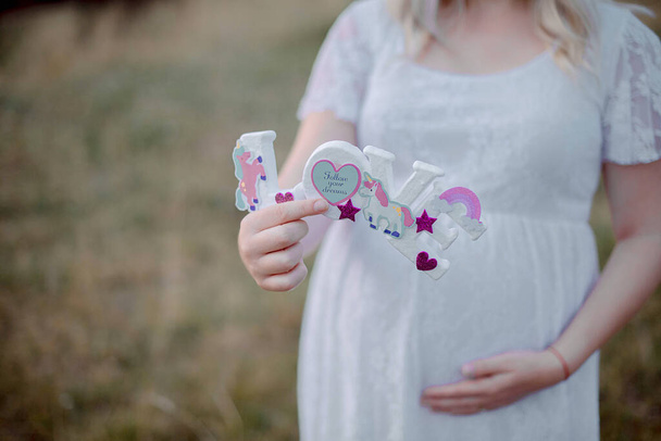 Mujer embarazada con un vestido de encaje blanco y mostrando orgullosamente su protuberancia bebé mientras sostiene un signo de letras de amor, esperando que la madre muestre felicidad con una decoración linda con estrellas, corazones y unicornios - Foto, imagen
