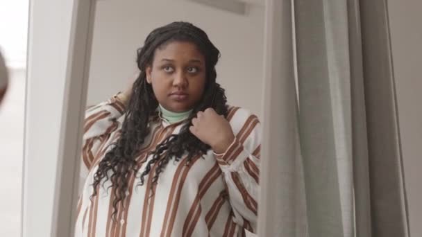 Hátsó kilátás a fiatal vonzó afro-amerikai nő visel alkalmi ruhák előtt áll a nagy tükör otthon, és simogatja a haját - Felvétel, videó